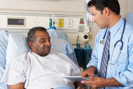 Clinician standing beside a patient in a hospital bed, reviewing information on a clipboard/tablet.